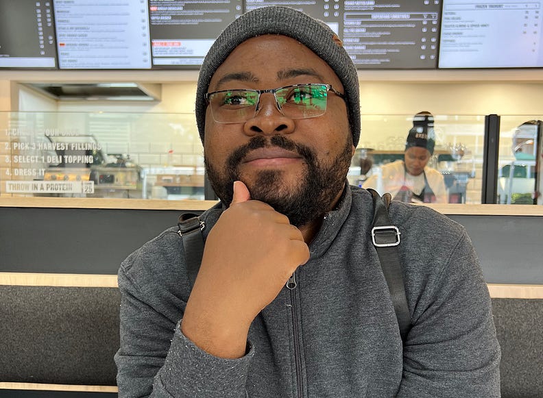 Photo of a man with a beard and glasses, wearing a hat and sweater, sitting in a cafe with his hand resting under his chin.