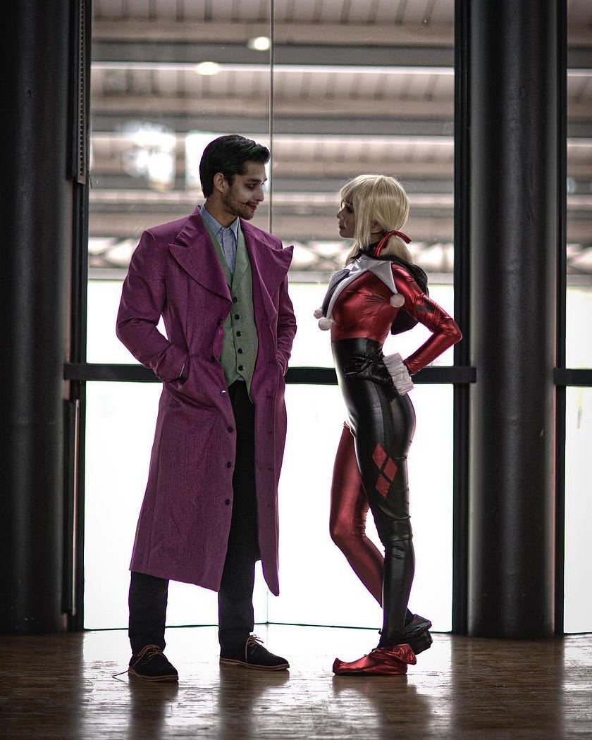 A man dressed up in purple trench coat portraying as Joker, stands next to a girl in yellow wig and a joker tightsuit, potraying as Harley Quinn