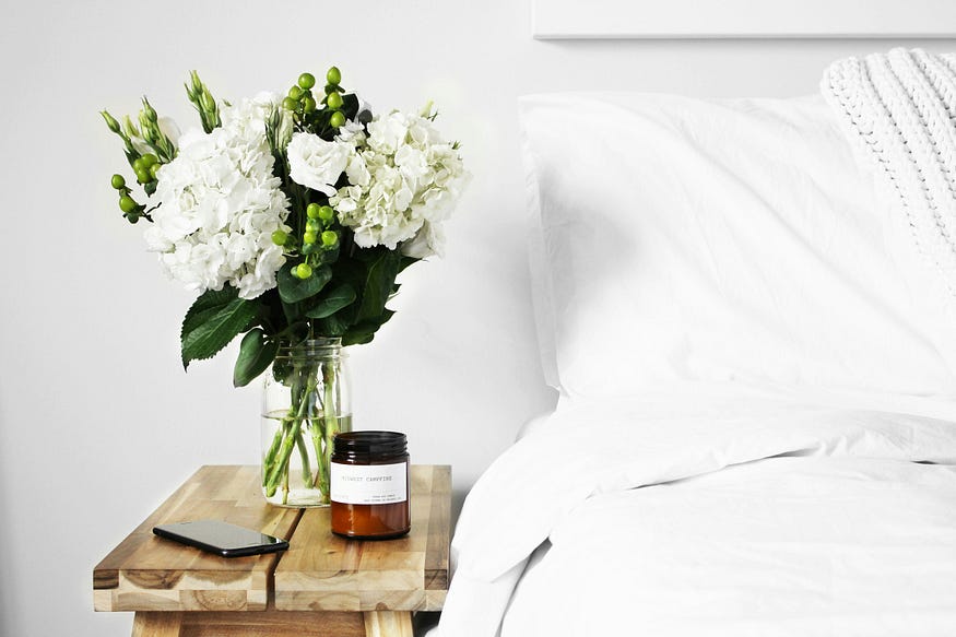 A bunch of fresh white carnations displayed on a bedstand next to a bed with white bed linen.