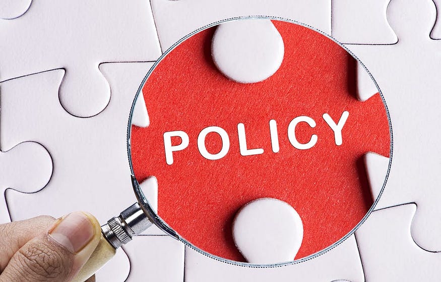 A magnifying glass is held over a red puzzle piece with the word POLICY written on it with a white puzzle pieces background; category creator, Carolyne Mweberi