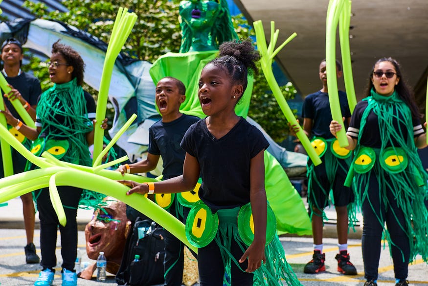 Children in Parade the Circle wearing green costumes and waving green celery-like constructions as they open their mouths to shout out.
