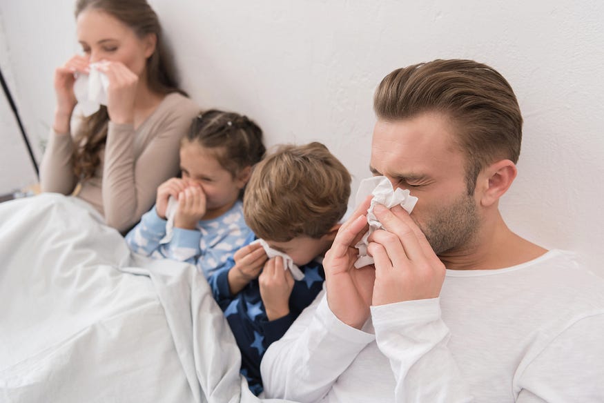 A father, mother, and two children are sitting in bed, blowing their noses from a bad cold.