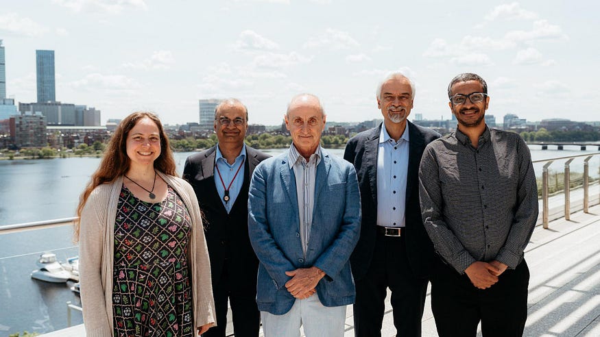 Physics colleagues pose with Peter Dourmashkin at the luncheon, with a view of the Charles River in the background.