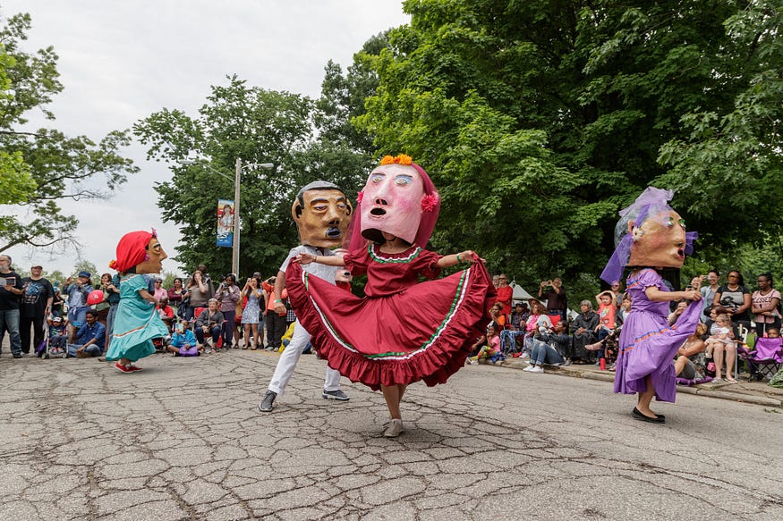 People in Parade the Circle dancing in brightly colored dresses wearing oversized papier-mâché heads. 