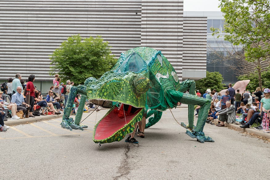 Crafted chameleon in Parade the Circle, taller than a person and operated by people inside the body, only their legs visible sticking out underneath. The chameleon has green skin and an open mouth, red inside.