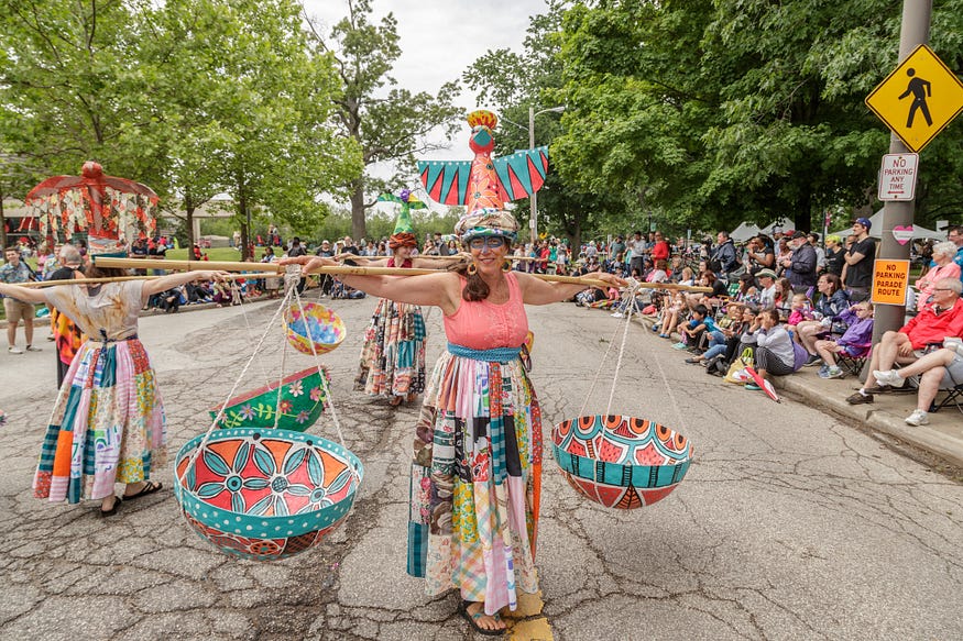 People walking in Parade the Circle with wooden poles draped over their shoulders and hung with a colorful basket on each side. Viewers gather on the sidewalks on either side.
