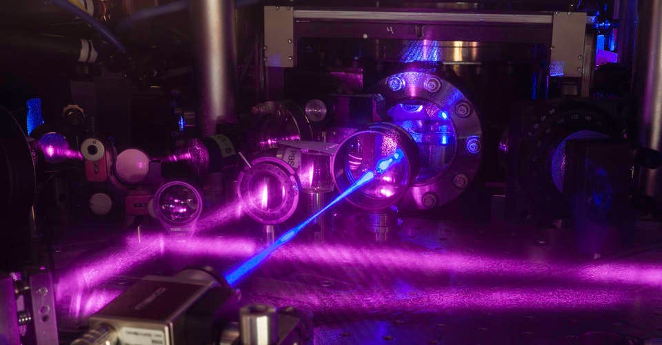 JILA’s three-dimensional (3-D) quantum gas atomic clock consists of a grid of light formed by three pairs of laser beams. A stack of two tables is used to configure optical components around a vacuum chamber. Shown here is the upper table, where lenses and other optics are mounted. A blue laser beam excites a cube-shaped cloud of strontium atoms located behind the round window in the middle of the table. Strontium atoms fluorescence strongly when excited with blue light. (Credit: G.E. Marti/JILA)