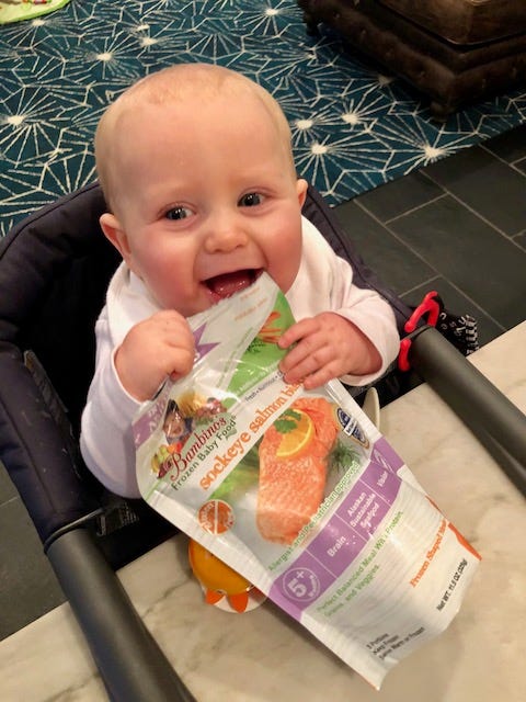 A cheerful baby in a high chair playfully chomping on a Bambino’s Frozen Baby Food package of sockeye salmon, showcasing early excitement for healthy eating and Bambino’s tasty offerings