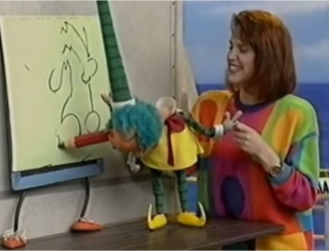 Scene from Mr Squiggle, a children’s TV show where a colourful puppet with blue hair is holding a red crayon and drawing on a large pad. A smiling woman wearing a bright, multicoloured sweater watches and guides the puppet.