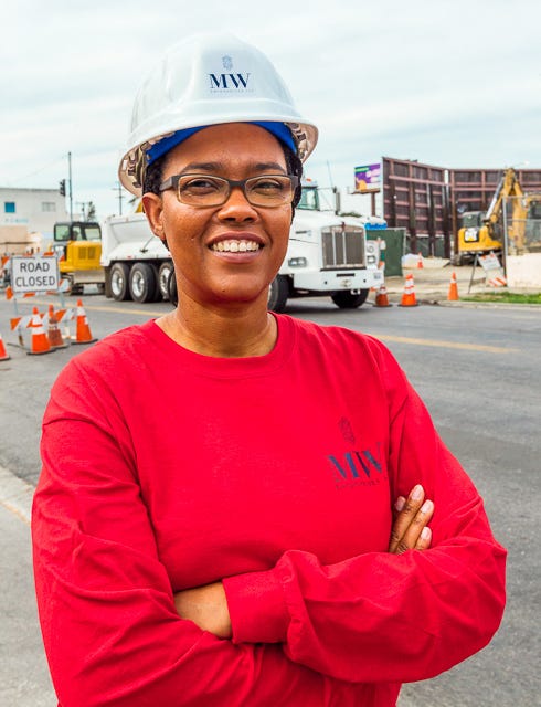 Mel Wyatt wears an MWE-branded white hard hat and MWE red longsleeve shirt with her arms crossed. Smiling, she observes a project site in Los Angeles.